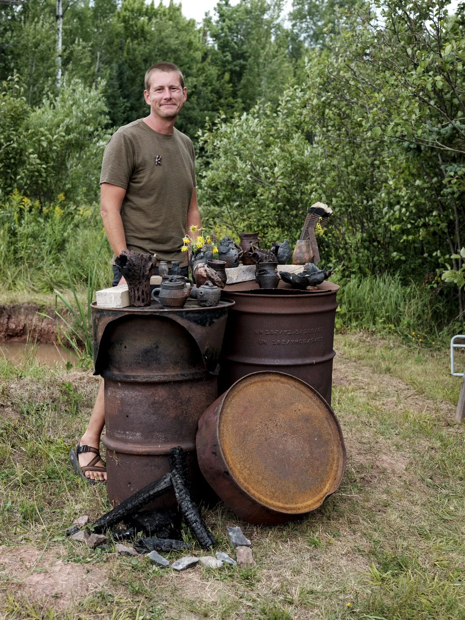 Richard Ward standing in a forest clearing behind a group of dark vessels on rusted metal drums