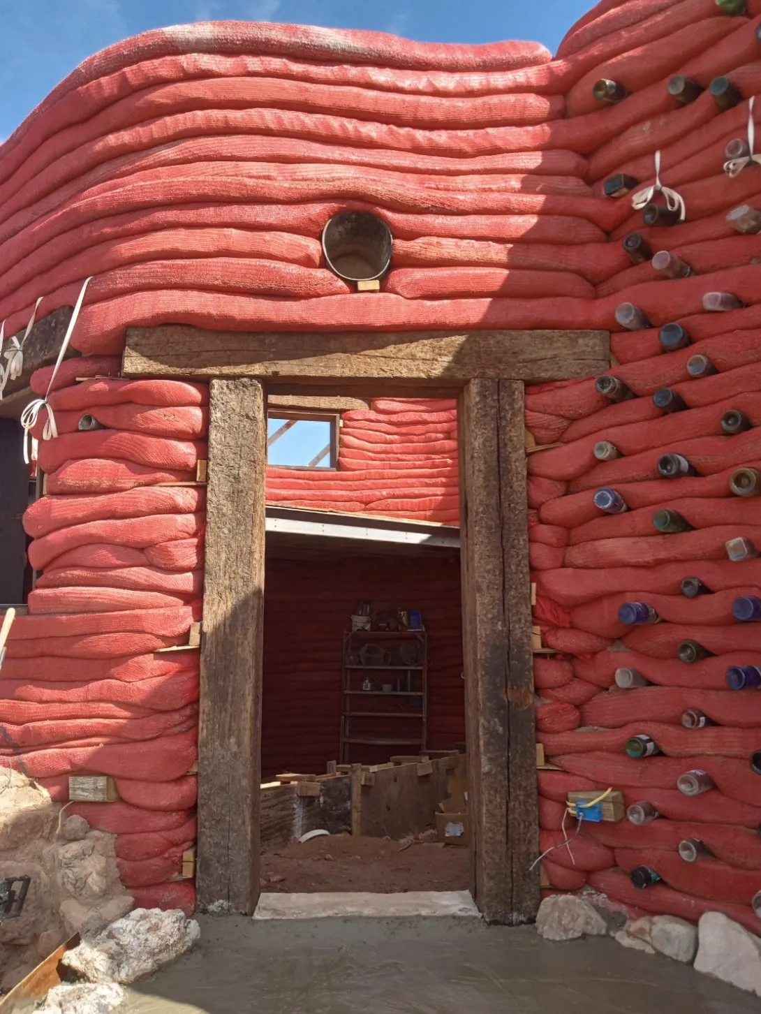 The earthbag home’s red plastered wall and wooden doorway, with bottles set into the wall