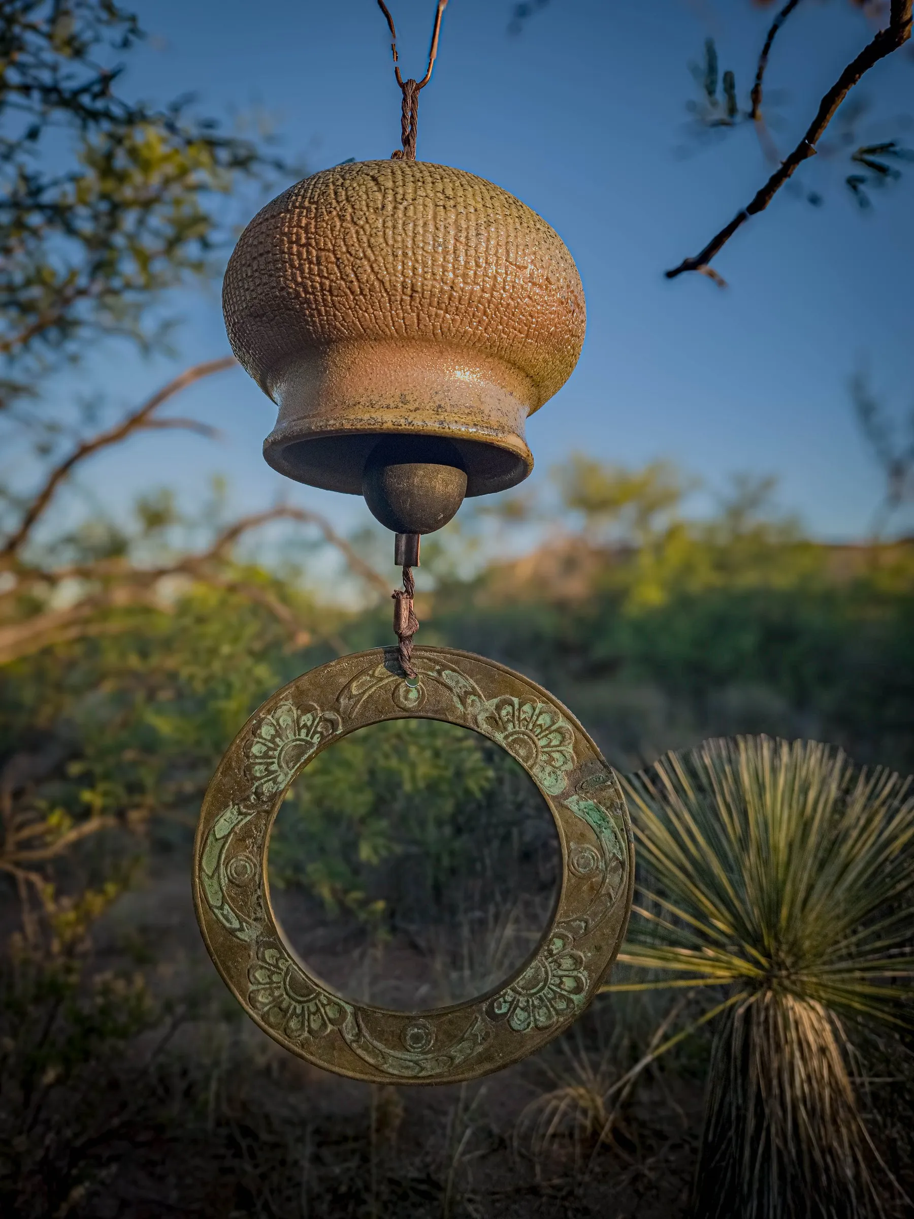 A wood-fired clay bell hanging above a brass ring among desert plants