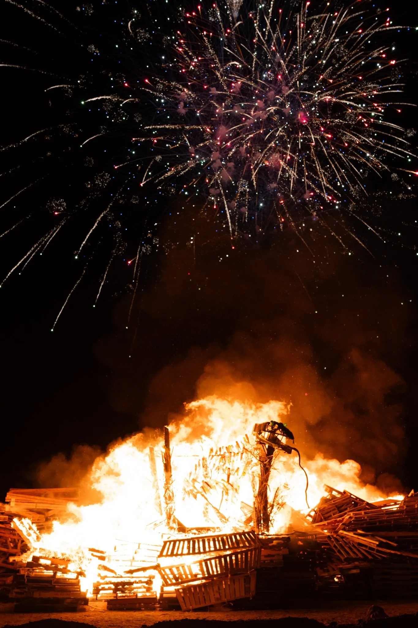 The rattlesnake sculpture burning on its pallet stack at night under fireworks