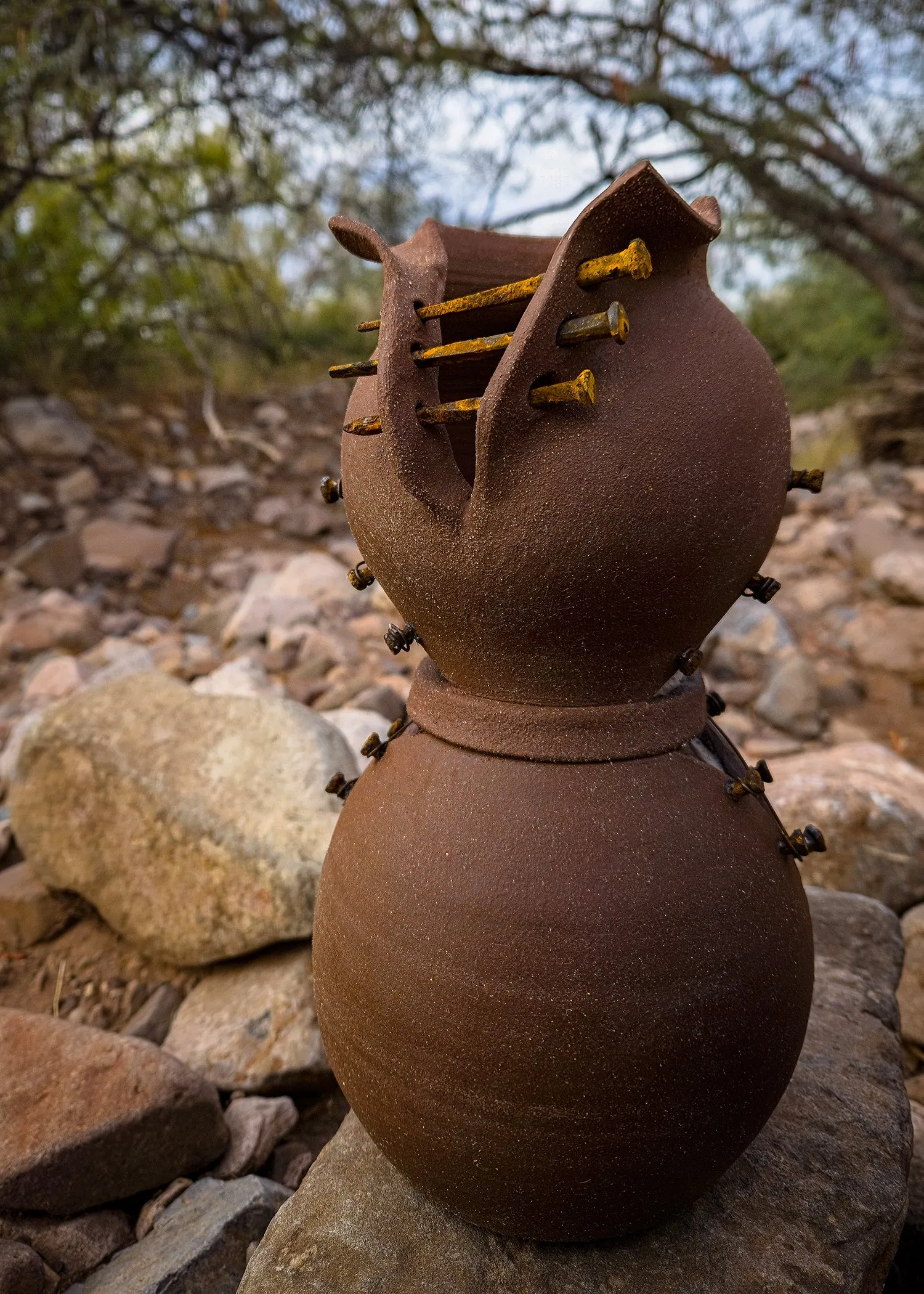 A stacked clay sculpture pierced with rusted nails, on a rock outdoors