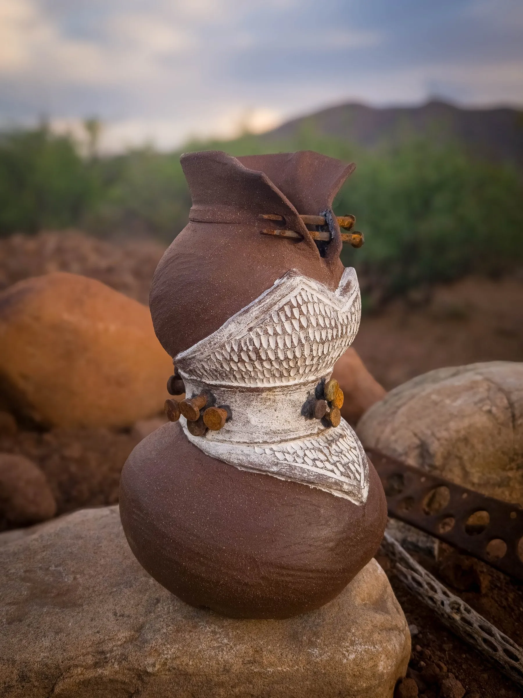 A stacked clay sculpture with a band of white lace texture and rusted nails, outdoors at dusk