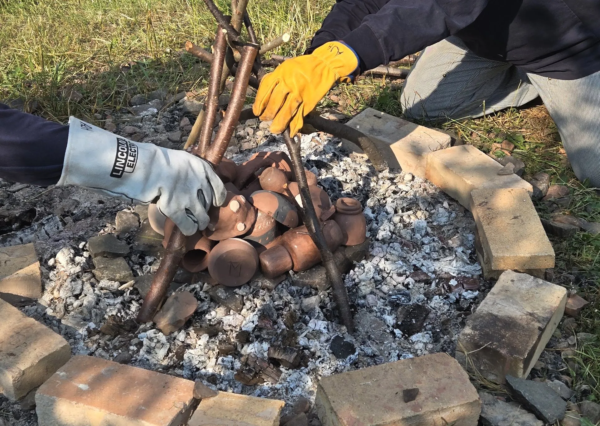 Gloved hands lifting pots out of a pit firing with long tongs