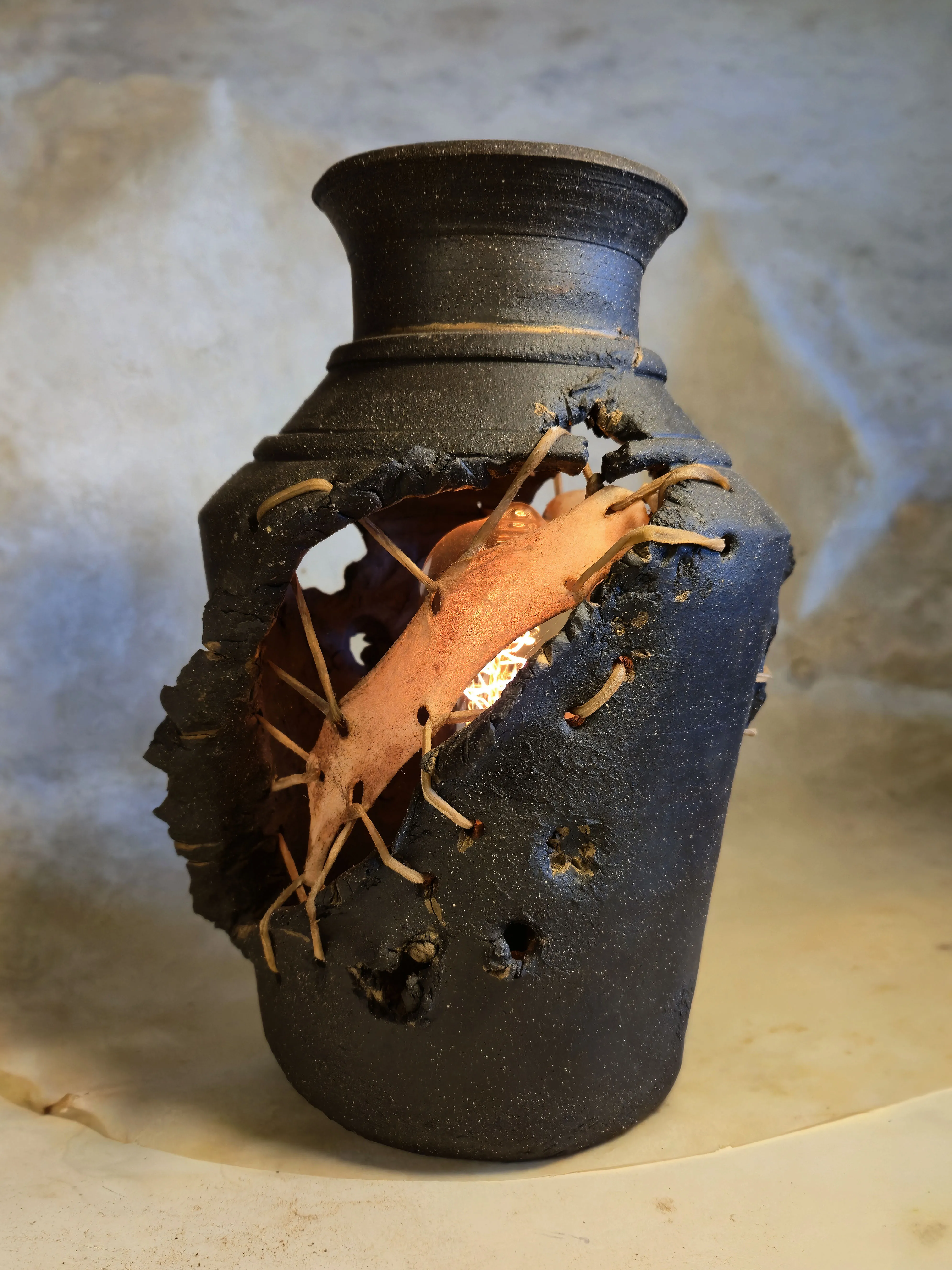 A tall black vessel torn open to show a lit Edison bulb glowing behind laced rawhide sheeting.